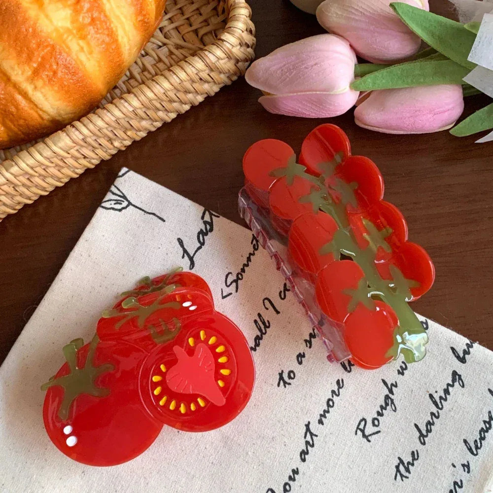 A flat lay arrangement showcasing two tomato-themed hair accessories on a textured white cloth, with a croissant in the background.