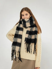 Woman wearing a cream cable knit sweater and a large black and beige plaid scarf with fringe, smiling and looking at the camera.