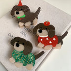 A collection of three festive acrylic dachshund dog brooches displayed on a neutral background. One wears a green sweater, another a red sweater with stars, and the third a red party hat.