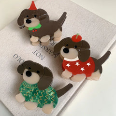 A collection of three festive acrylic dachshund dog brooches displayed on a neutral background. One wears a green sweater, another a red sweater with stars, and the third a red party hat.