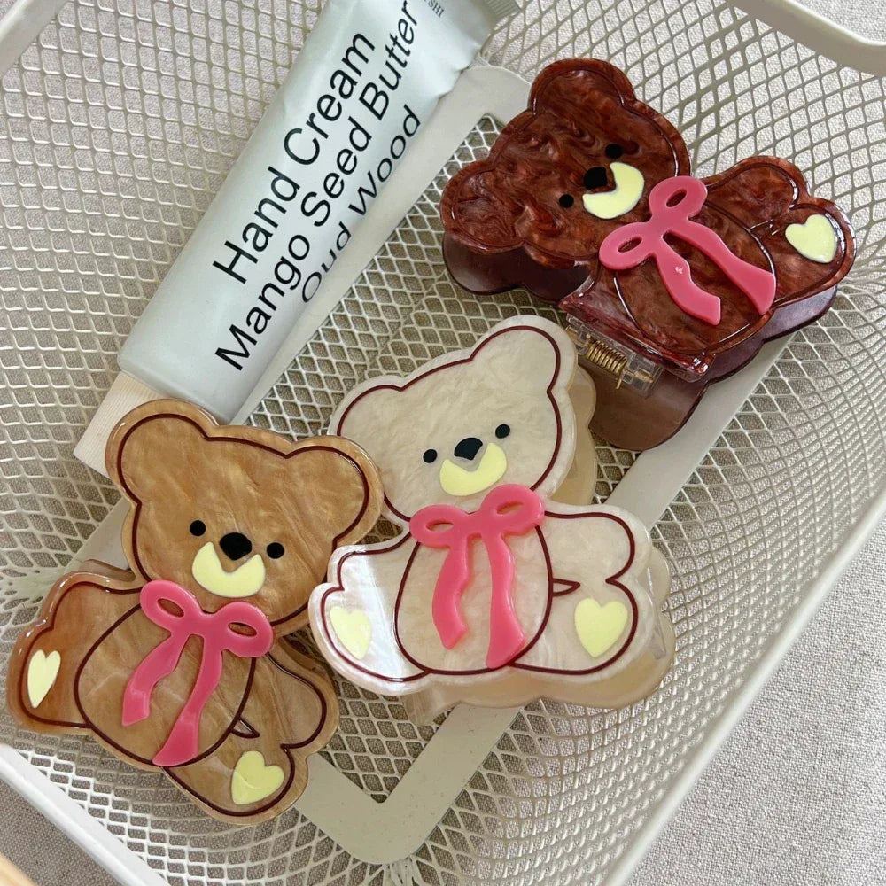Three teddy bear hair clips in brown, beige, and cream colors, arranged with a tube of hand cream in a white basket.