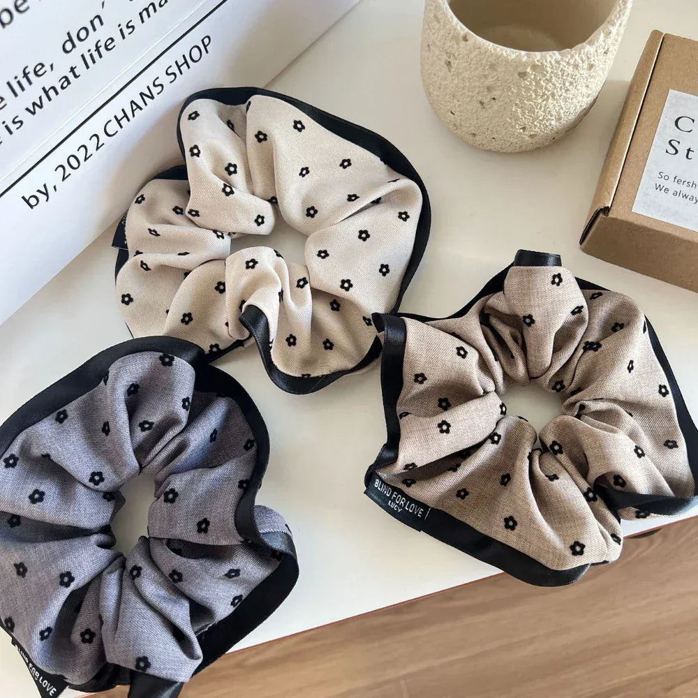 Three scrunchies arranged on a white surface, one beige, one grey, and one tan, all with black floral prints and black trims.