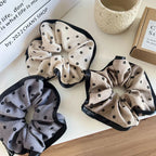 Three scrunchies arranged on a white surface, one beige, one grey, and one tan, all with black floral prints and black trims.