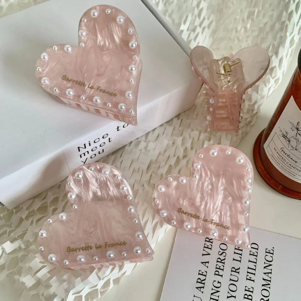 Four pink marbled heart-shaped hair clips with faux pearl embellishments, displayed on a white surface with decorative elements and a candle.