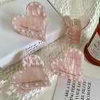 Four pink marbled heart-shaped hair clips with faux pearl embellishments, displayed on a white surface with decorative elements and a candle.