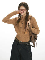 Woman wearing a brown cable knit hooded sweater with buttons and pom-poms, dark wash wide-leg cropped jeans, a black belt, and a brown crossbody bag, smiling and touching her hair.