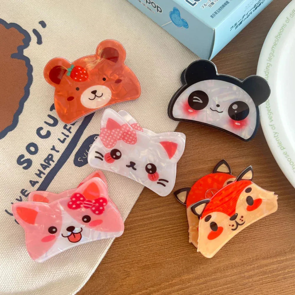 A flat lay of various cute animal-shaped hair clips including a bear, panda, cat, corgi, and fox, arranged on a white cloth and wooden surface.