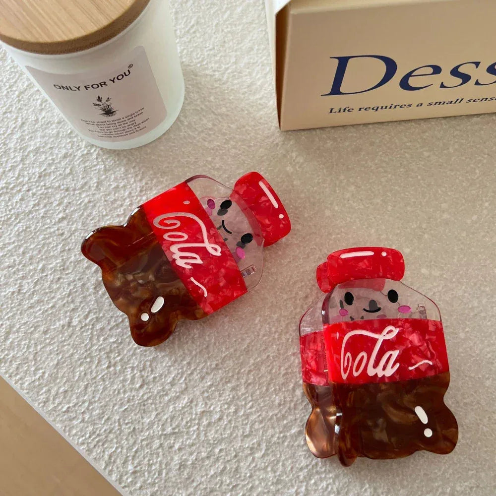 Two cute cola bottle hair clips are displayed on a white surface with a candle and a box.