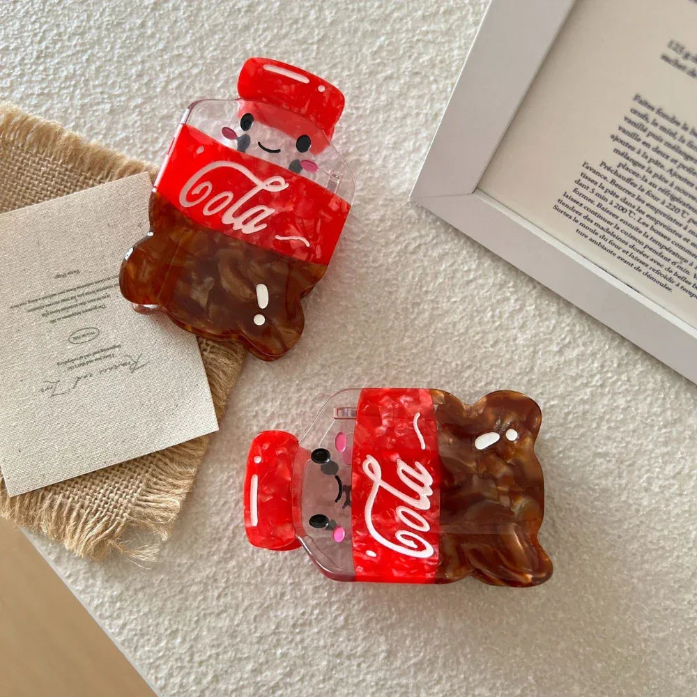 Two cute cola bottle hair clips are shown on a white surface next to a framed picture and a card.