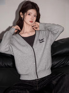 Woman wearing a grey knit cardigan with black trim and embroidery, layered over a dark top, paired with dark wide-leg jeans, sitting on a black couch.