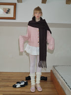 A young woman poses with her arms outstretched slightly, wearing a pink knit cardigan over a white pleated skirt, a long dark scarf, white tights, leg warmers, and pink ballet flats.