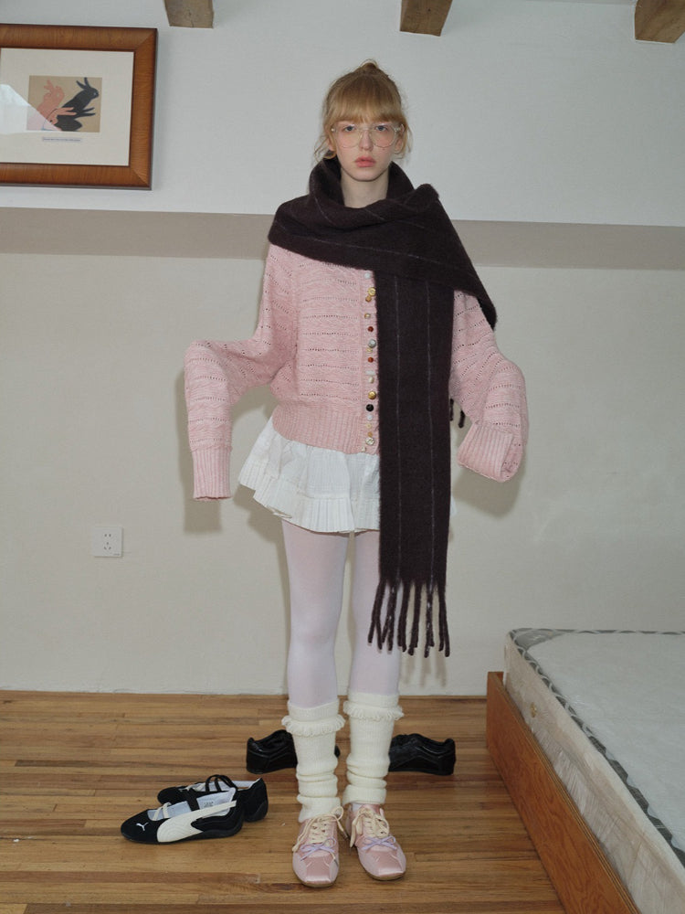 A young woman poses with her arms outstretched slightly, wearing a pink knit cardigan over a white pleated skirt, a long dark scarf, white tights, leg warmers, and pink ballet flats.