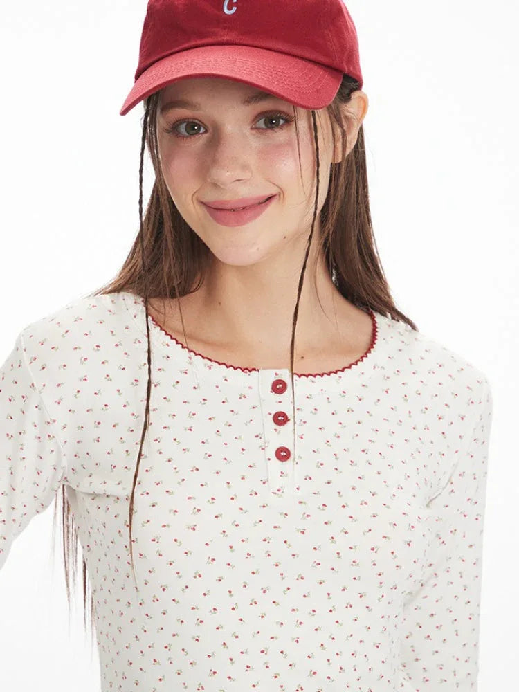 Front view close-up of a model's face and upper body, wearing a white long-sleeve top with a floral print and red trim, and a red baseball cap.