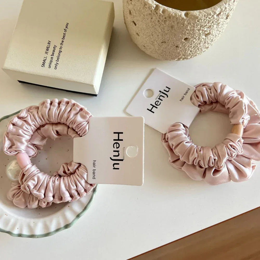 Two light pink satin scrunchies with ruffled texture are arranged on a white surface. One scrunchie features a wooden bead, while the other is adorned with a pearl.