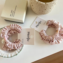 Two light pink satin scrunchies with ruffled texture are arranged on a white surface. One scrunchie features a wooden bead, while the other is adorned with a pearl.