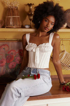 Woman wearing a white ruched corset top with a bow accent and light wash denim jeans, sitting on a counter.
