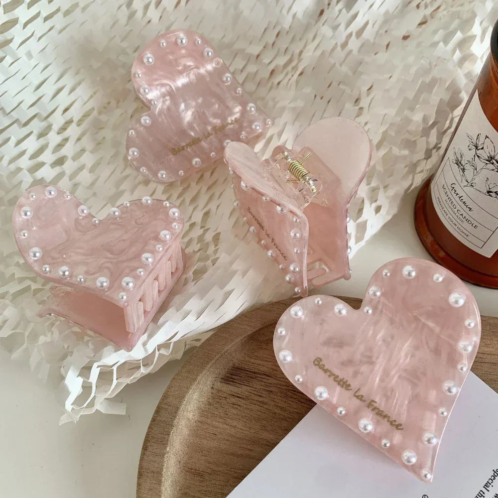 A close-up of four pink marbled heart-shaped hair clips with faux pearl embellishments, arranged on a wooden tray and beside a scented candle.