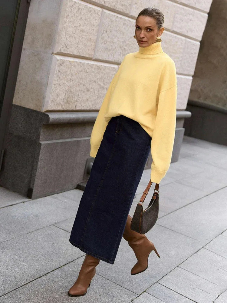 Woman wearing a yellow oversized turtleneck sweater with a dark denim maxi skirt, brown boots, and a brown handbag, walking outdoors.