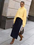 Woman wearing a yellow oversized turtleneck sweater with a dark denim maxi skirt, brown boots, and a brown handbag, walking outdoors.