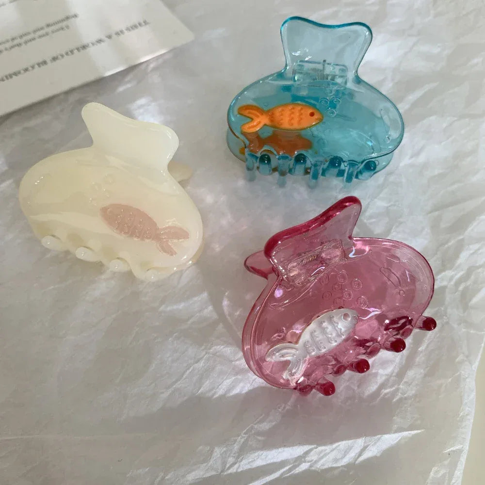 Three hair claw clips in assorted colors (cream, blue, pink) with fish charms, arranged on white paper.