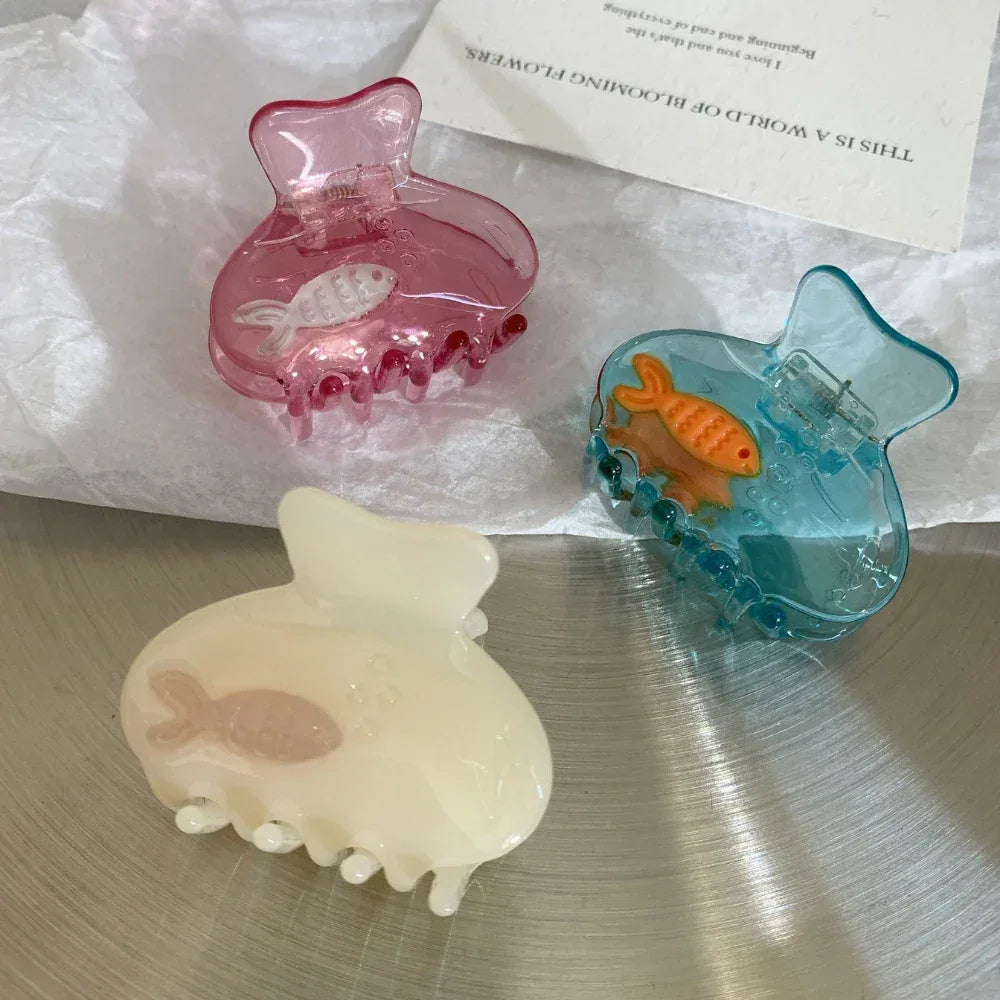 A trio of decorative hair clips in pink, blue, and cream, each containing a small fish.