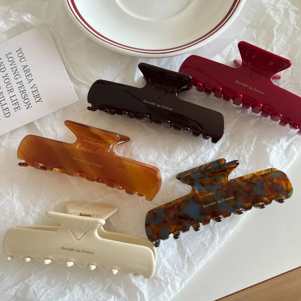 An overhead shot showcasing five hair claw clips in red, dark brown, amber, tortoiseshell, and cream, placed on a white surface with scattered white paper.