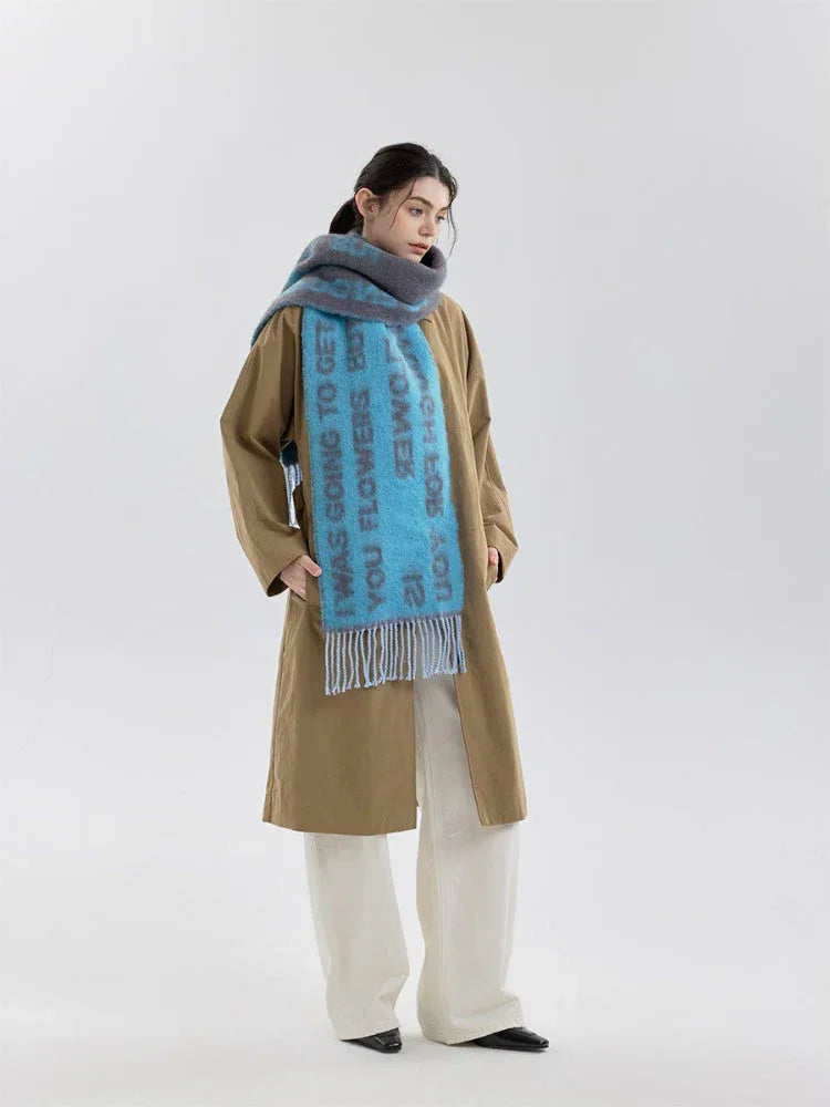 A woman models a blue and gray knit scarf with text print and fringe detail, worn over a tan trench coat.