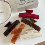 A group of five hair claw clips in red, dark brown, amber, tortoiseshell, and cream colors, artfully arranged on a white background.