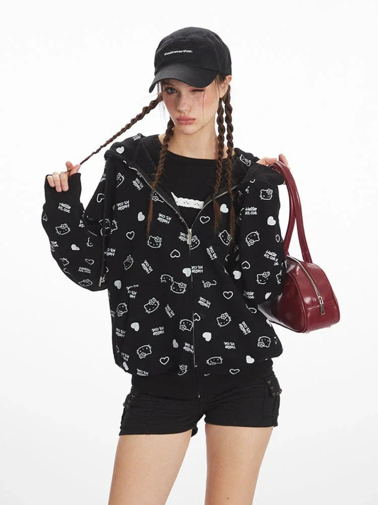 Close-up front view of a model wearing a black zip-up hoodie with an all-over white Hello Kitty and heart graphic print, black t-shirt, black shorts, black knee-high socks, and red sneakers. The model is wearing a black baseball cap and has her hair in braids.