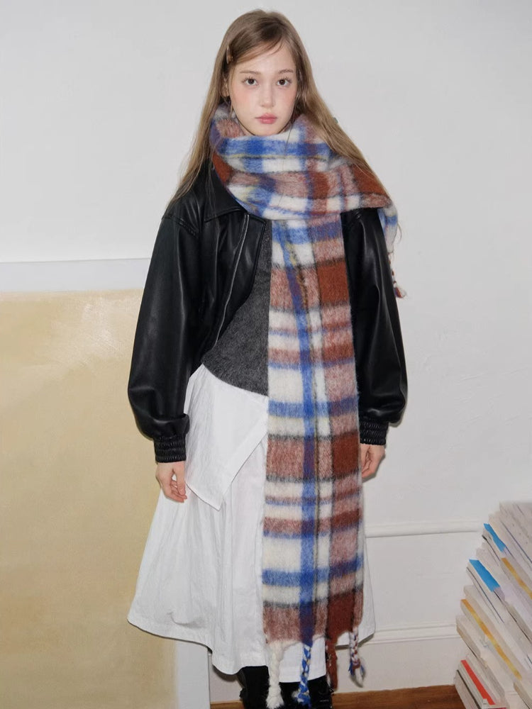 Model looking directly at the camera, wearing a black jacket, grey sweater, white skirt, and a prominent plaid scarf.