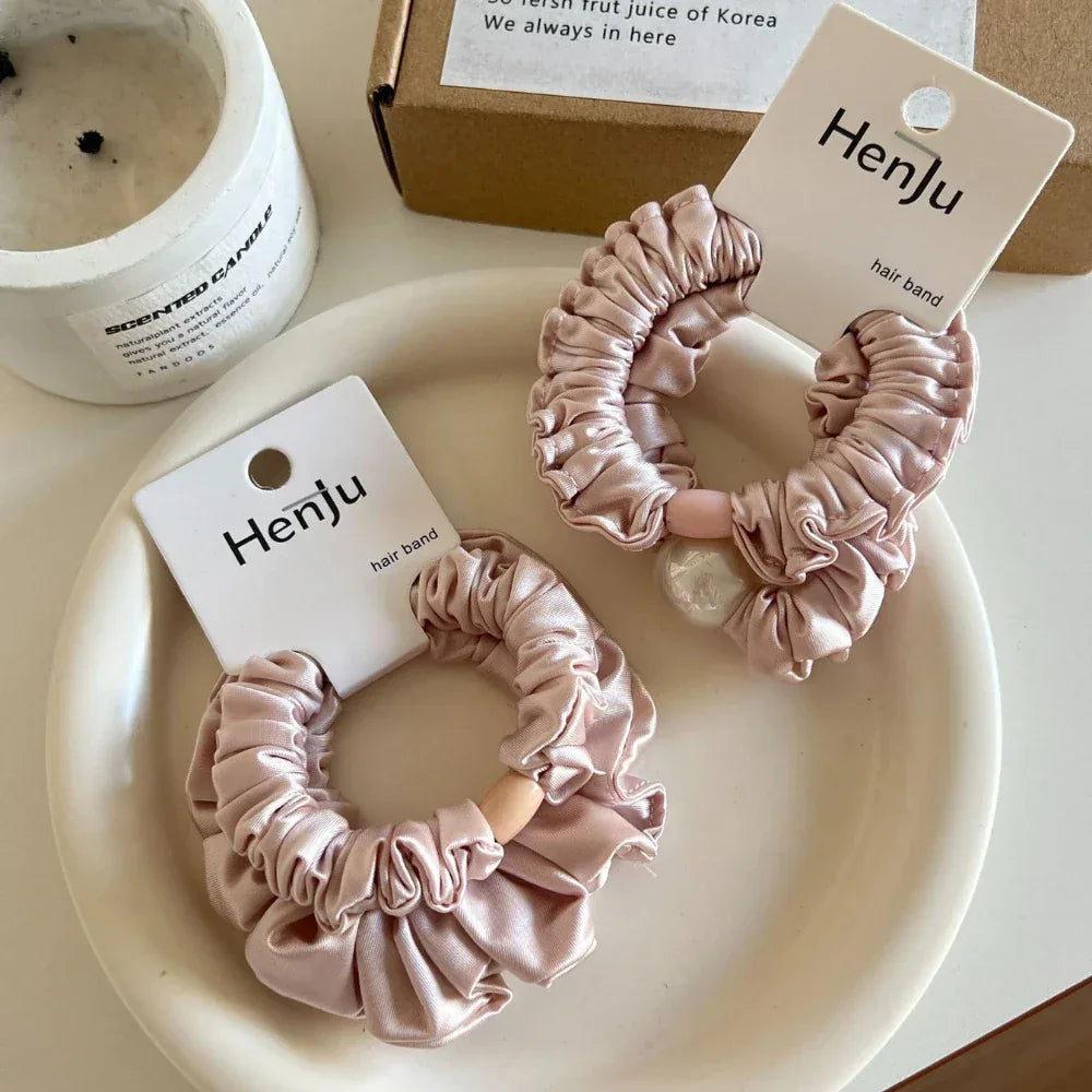 Two light pink satin scrunchies with ruffled texture are shown on a white plate. One scrunchie has a wooden bead, and the other is decorated with a pearl.