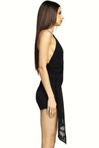 Side view of a woman wearing a black halter neck mini dress with a deep V-neckline and an asymmetrical hemline with a sheer wrap-around skirt detail. The dress features side cut-outs and an open back.