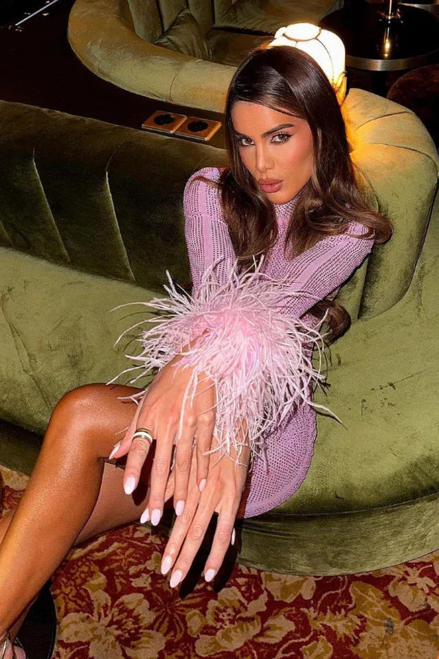 Woman with dark hair sitting on a green couch wearing a pink long-sleeved dress with feather cuffs.