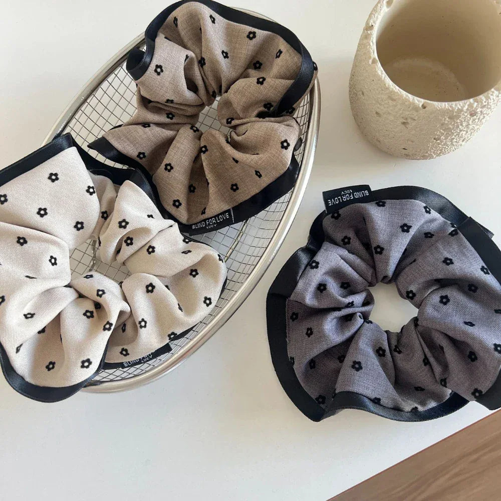 A metal basket containing two scrunchies with floral patterns, and a third scrunchie with a floral pattern placed beside it on a white surface.