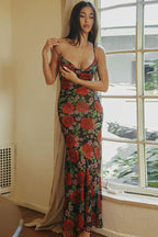 Woman wearing a long floral print dress with spaghetti straps and a cowl neckline, standing by a window.