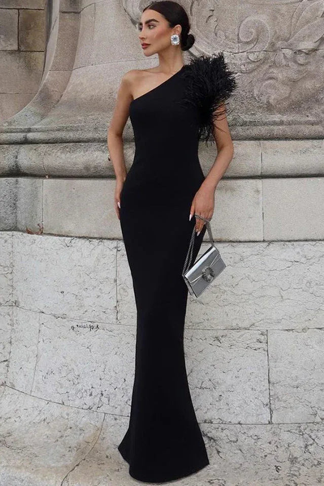 Woman wearing a black one-shoulder crepe gown with a feather accent on the shoulder and a mermaid hemline, standing in front of a stone wall. Main image