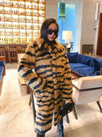 Woman in a faux fur tiger stripe coat and black boots, holding a black handbag, standing in a lounge area.