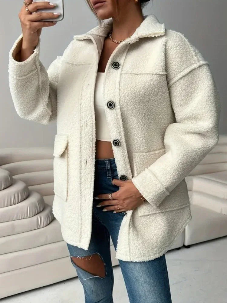 Woman holding her phone and wearing an oversized white sherpa fleece jacket over a white crop top and blue jeans.
