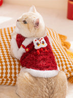 A cream-colored cat wearing a red and white festive vest with a lion dance embellishment, resting on a cushion.