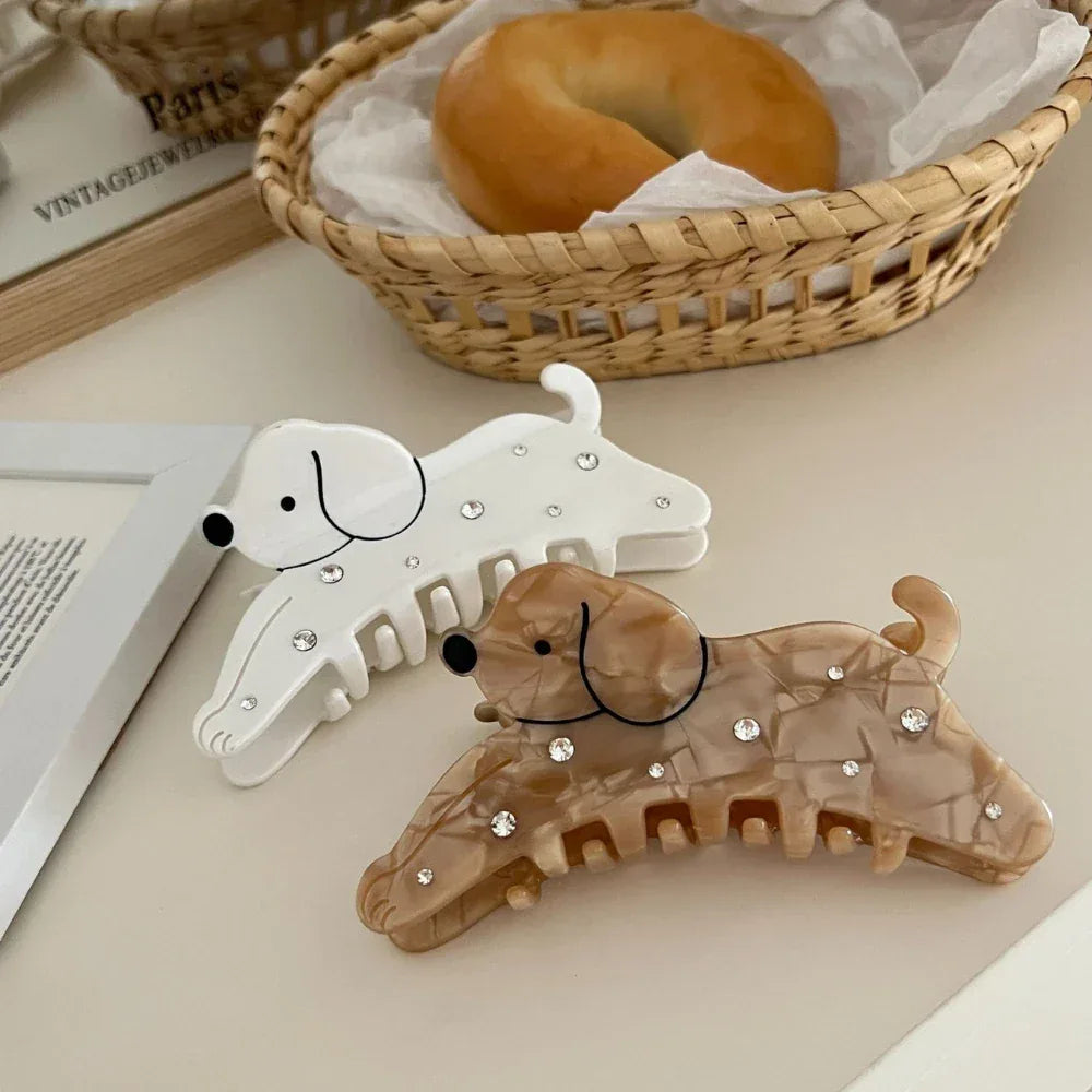 Two dog-shaped hair claw clips, one white and one brown, with sparkling embellishments, placed on a surface near a basket of bagels