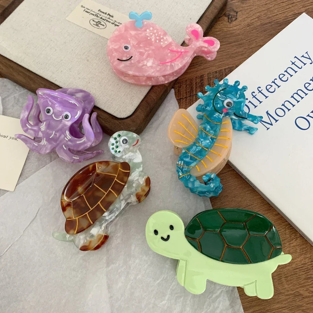 A flat lay of acrylic hair clips featuring a pink whale, a purple octopus, a blue seahorse, and two turtles arranged on a wooden surface and white paper.