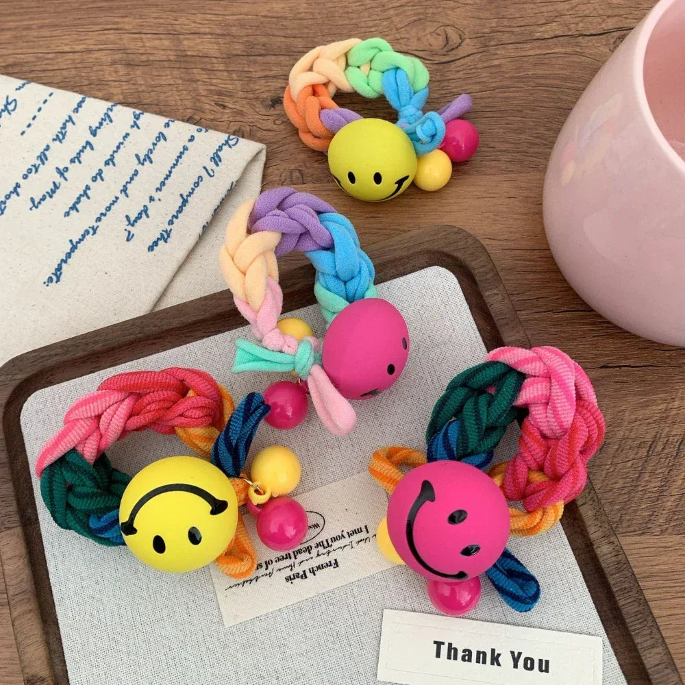 A close-up view of several playful braided hair ties, each adorned with a smiley face charm and accent beads, on a textured background.