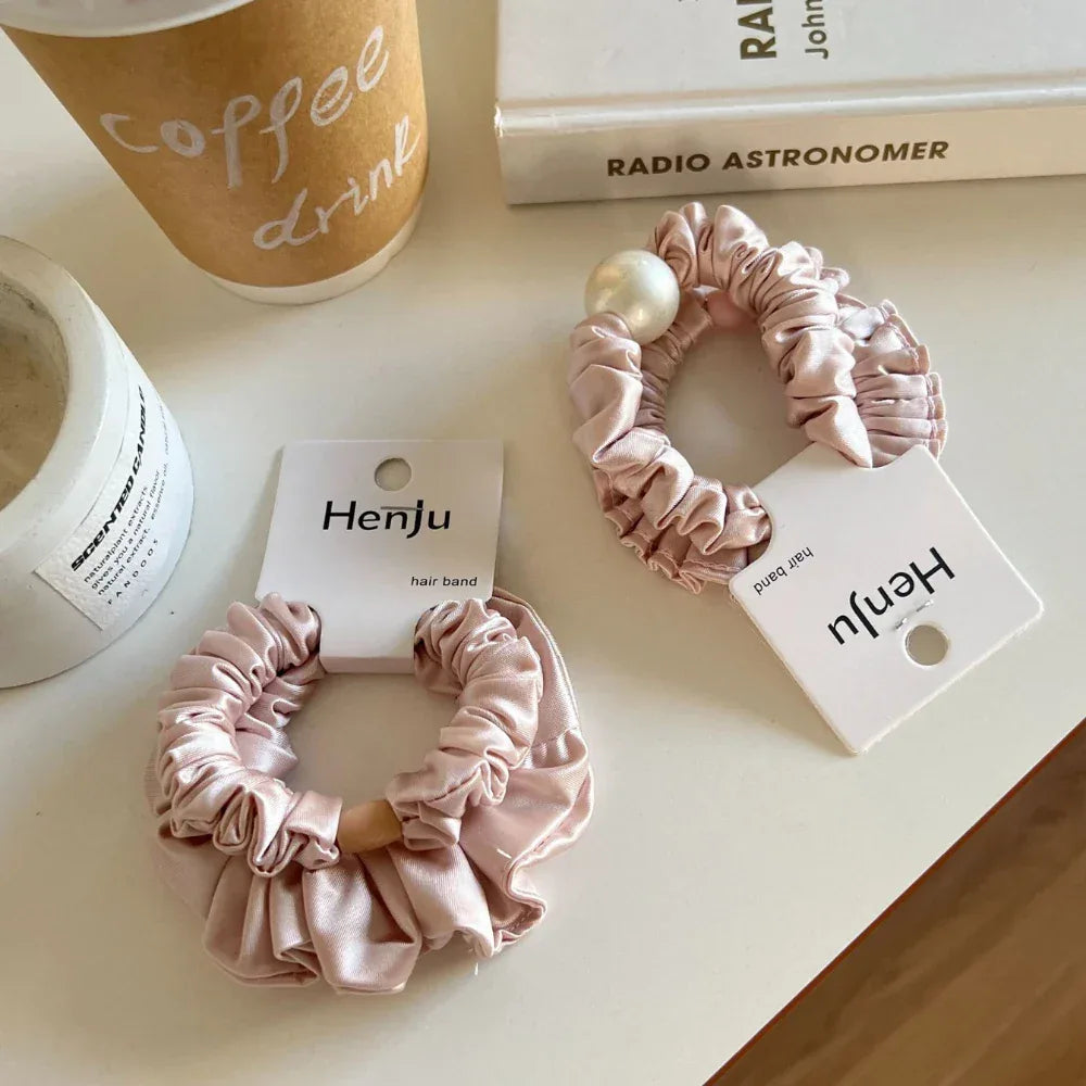Two light pink satin scrunchies with ruffled texture are placed on a white table. One scrunchie has a wooden bead, and the other is embellished with a pearl.