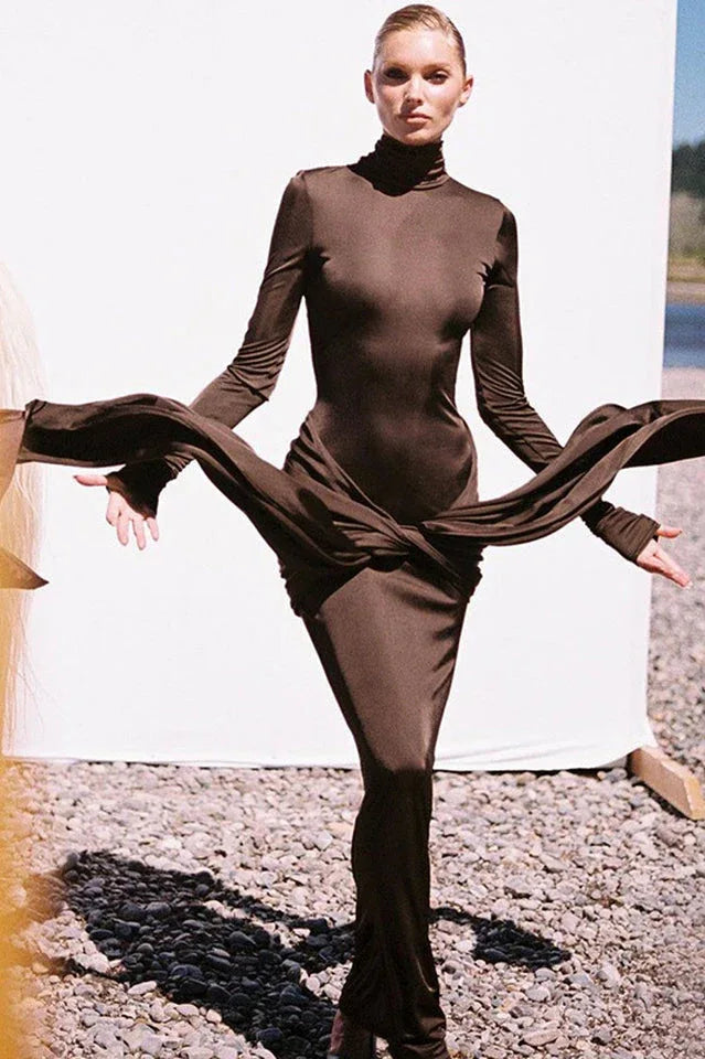 Woman wearing a long-sleeved, brown, mock neck, floor-length dress with a draped knot detail at the waist, with fabric draped around her arms.