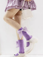 Full-body shot of a person wearing a purple ruffled skirt, white Mary Jane shoes, and purple socks adorned with large bows.