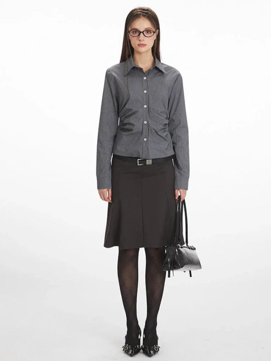 Woman wearing a grey long-sleeved button-down shirt with ruching detail, a black skirt, black tights, and black heels, holding a black handbag.