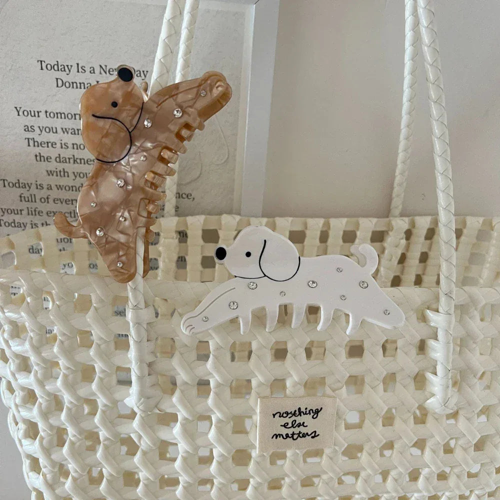 Two dog-shaped hair claw clips, one white and one brown, with sparkling embellishments, attached to a white woven bag