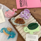 A close-up of various acrylic hair clips, including a pink whale, a purple octopus, a blue seahorse, and two turtles, set against a wooden background and white paper.