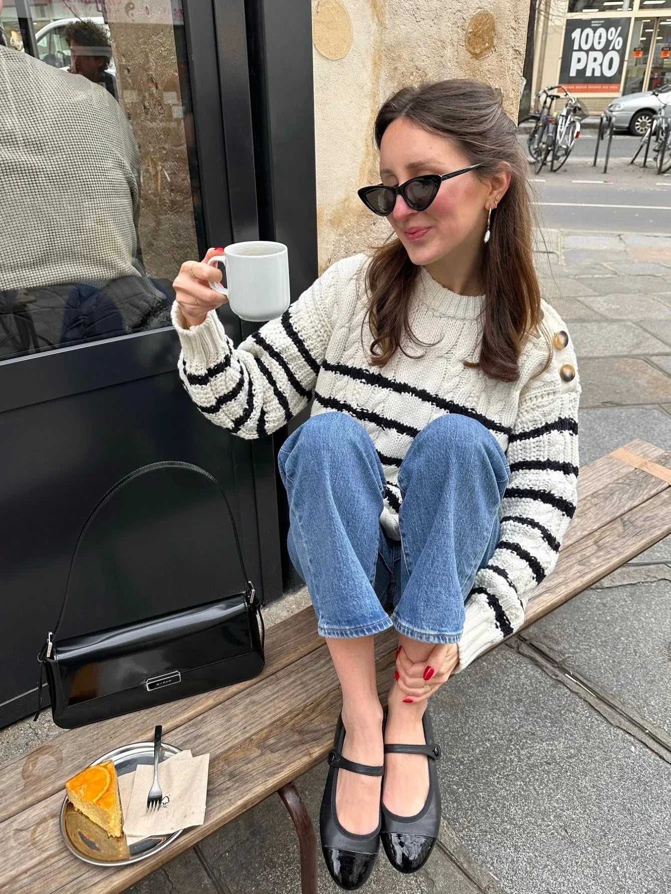 Woman sitting on a bench outside a cafe, holding a coffee cup and wearing a striped knit sweater and blue jeans.