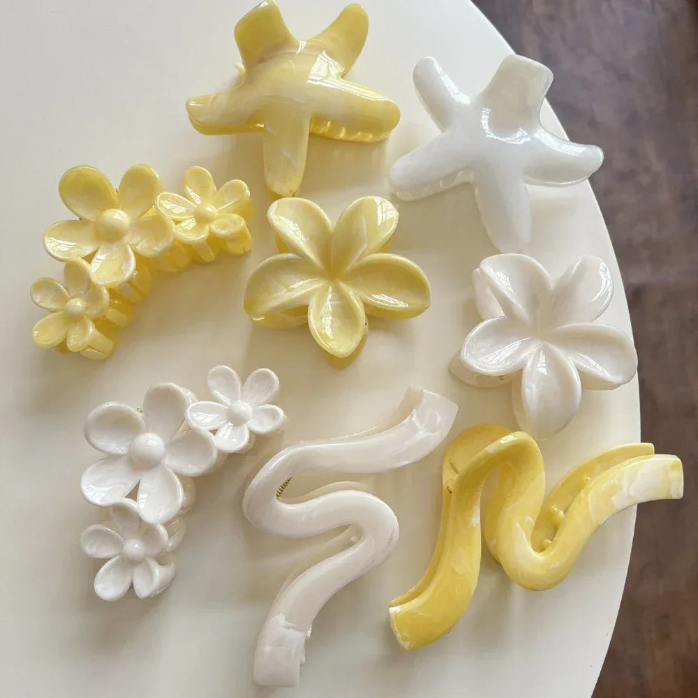 An assortment of yellow and white hair accessories, featuring floral designs, star shapes, and wavy claw clips, spread across a white surface